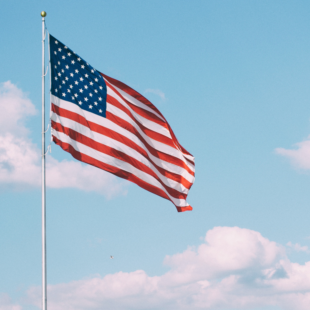 An American flag wraps around a flagpole as the sun shines through it beneath a wide, cloud-dotted blue sky.