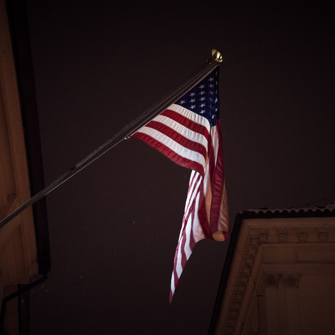 An American flag hangs from an angled flagpole as a light below illuminates it against the night sky.