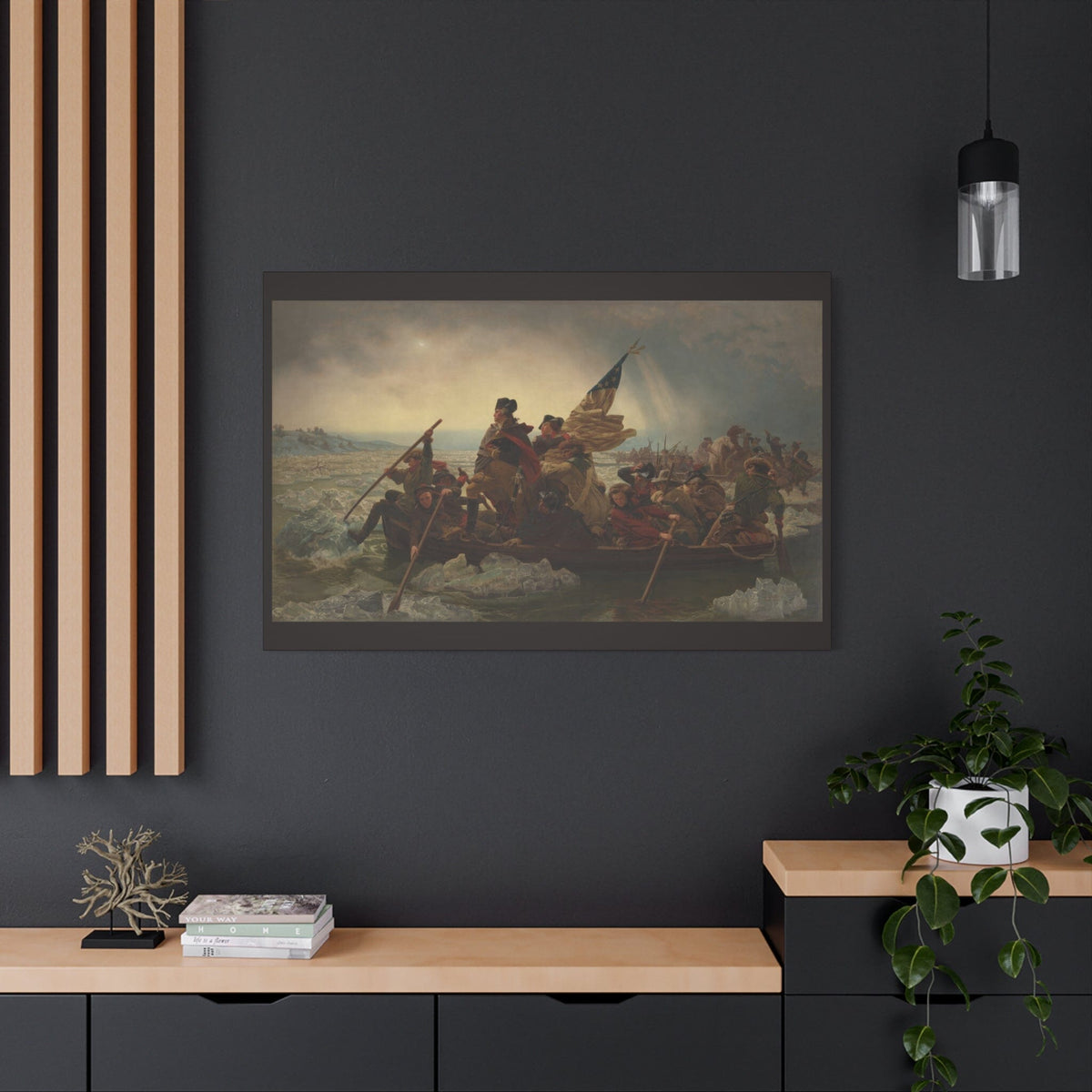 A framed historical painting of George Washington leading a Continental Army across a snowy battlefield, displayed on a dark charcoal wall above a light wood console table with a plant and decorative objects.