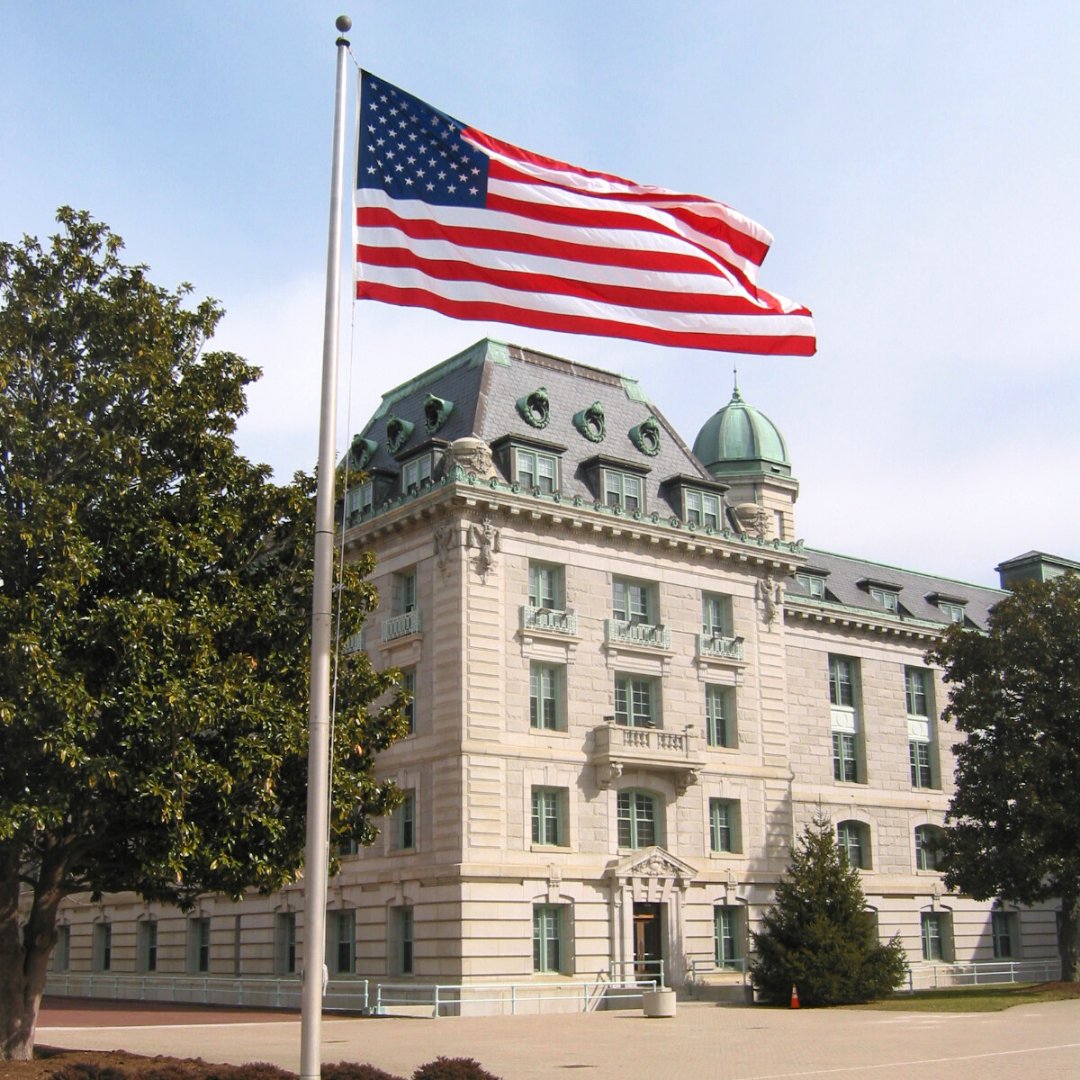 An American flag flies on a tall flagpole in front of a large stone campus building with trees and a green-accented roof.