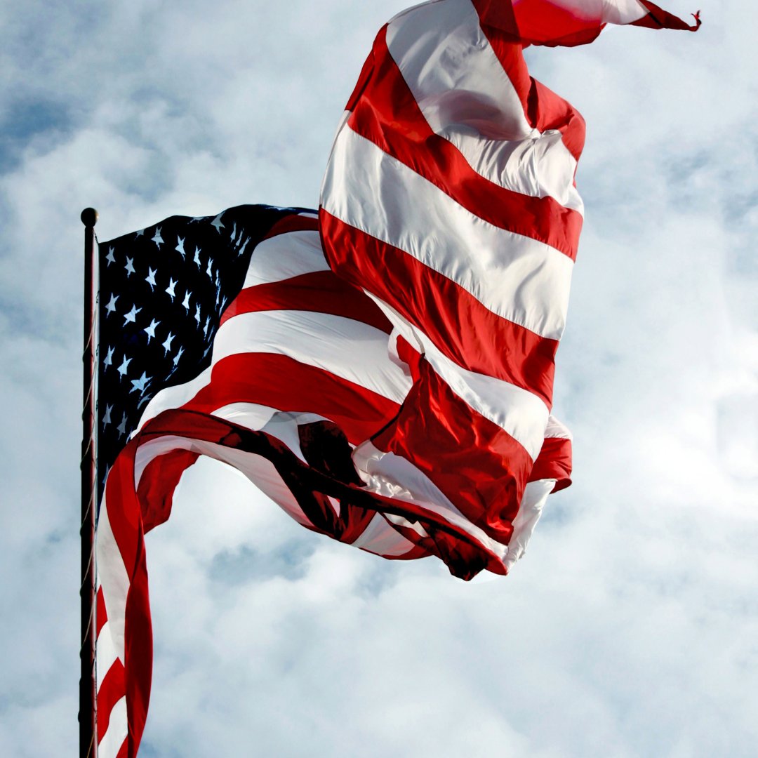 An American flag billows in strong wind beside a flagpole as the flag's red and white stripes fill a cloudy blue sky.