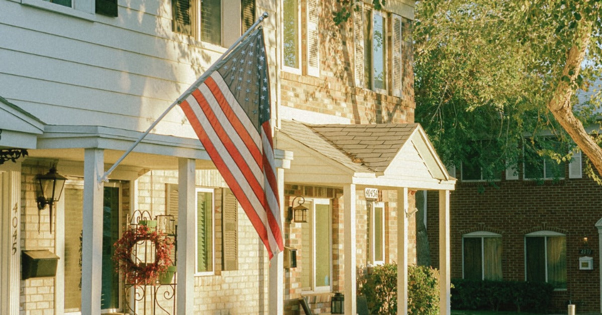 An American flag hanging from a house porch above a walkway, with neighboring brick homes and trees nearby.